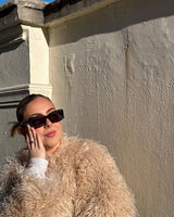 Woman in sunglasses and faux sheep jacket beige leaning against a wall.
